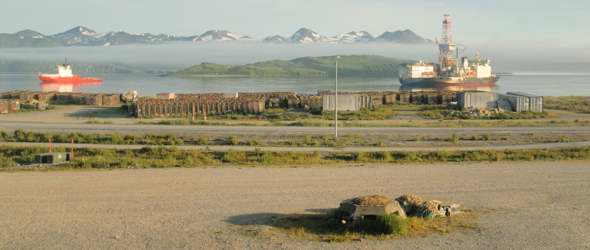 View of Unalaska Bay from Grand Aleutian Hotel, with concrete bunker in foreground, bike path and two-lane paved Airport Beach Road behind it, junk on the shore, and two ships anchored in the bay