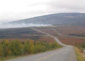 Paved portion of highway, with elevated pipeline on right dropping underground, and wildfire in the distance